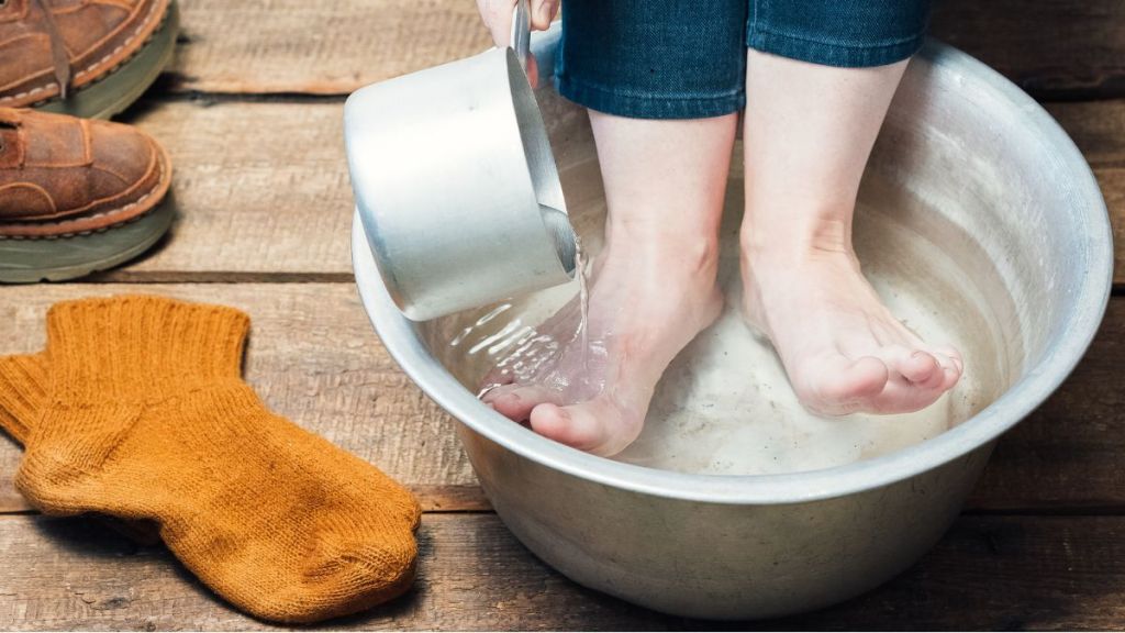 Pieds plongés dans une bassine d’eau fraîche, avec une main versant de l’eau, sur un sol en bois, à côté de chaussettes en laine orangée.