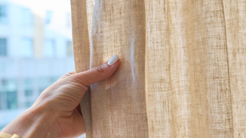 Main entrouvrant un rideau en lin beige, laissant filtrer la lumière extérieure d’un immeuble en arrière-plan.